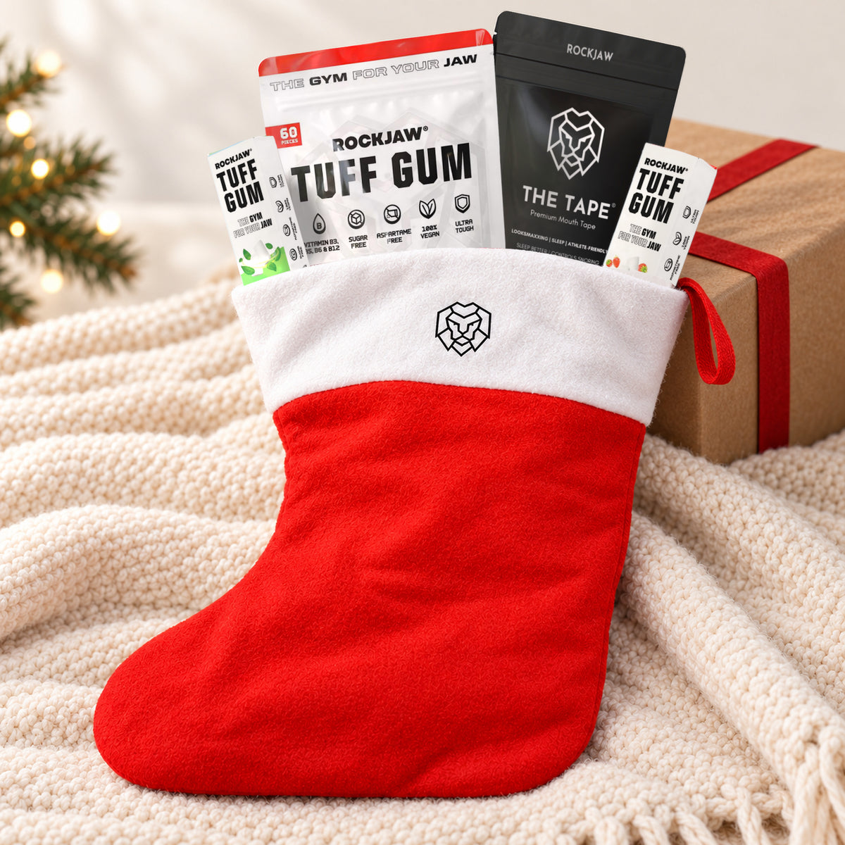 The ROCKJAW Stocking, a red felt Christmas stocking with a white cuff and the ROCKJAW lion logo, leaning against a kraft-wrapped present on a cream knitted throw, filled with a Tuff Gum Strawberry Smash 60 piece pouch, The Tape mouth tape and two small Tuff Gum packs.